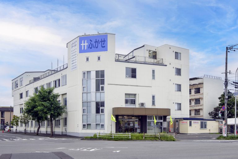 白色4階建ての介護施設建物。紫色の「けわせ」看板が目立つ。