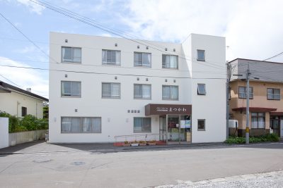 白色3階建ての介護施設の建物外観。玄関入口と駐車スペースが見える。